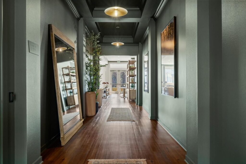 Hallway of clubhouse with large mirror, art, overhead lighting and various decor including bookshelves and plants
