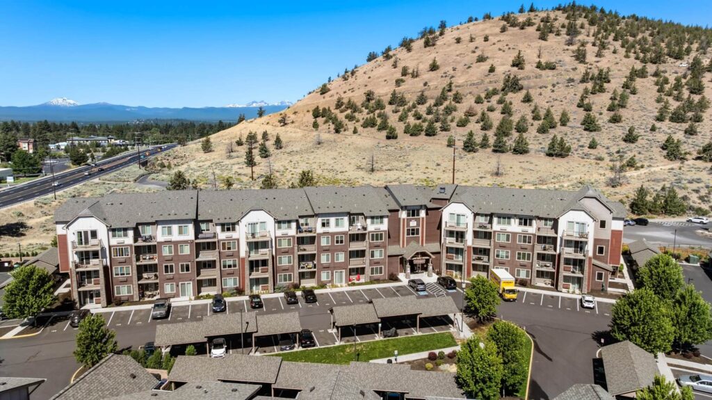 Exterior view of four story apartment building with parking lot and covered parking, with large hill behind it