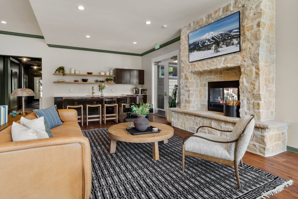 Clubhouse lounge room with sofa and armchair seating next to a fireplace with a television above it, next to a small kitchen space