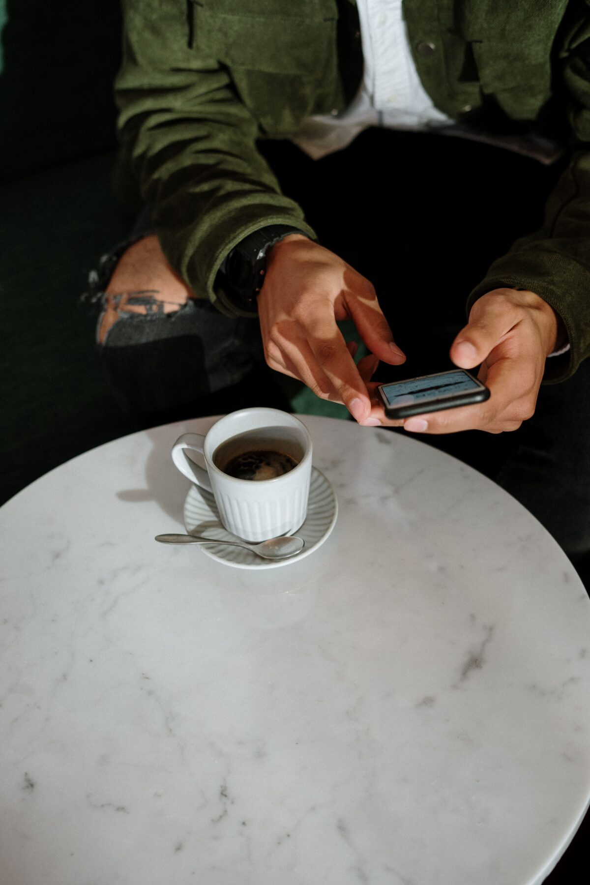 A person wearing a green jacket and black watch sits at a white marble table, holding a smartphone. A cup of coffee on a saucer with a spoon rests on the table. The persons face is not visible.